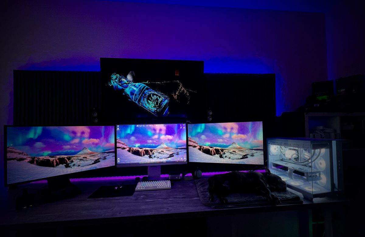 The Lab: triple-monitor desk in purple ambient light with a white water-cooled build and Tyson the Frenchie asleep on the desk